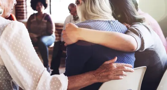 Two women embrace in a supportive group setting, with others seated in the background, fostering a sense of community.