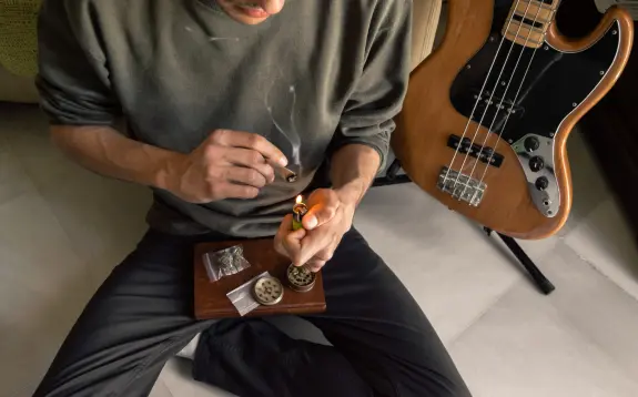 A person lights a joint while seated on the floor, with a bass guitar and cannabis supplies nearby.