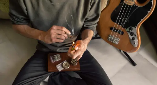A person lights a joint while seated on the floor, with a bass guitar and cannabis supplies nearby.
