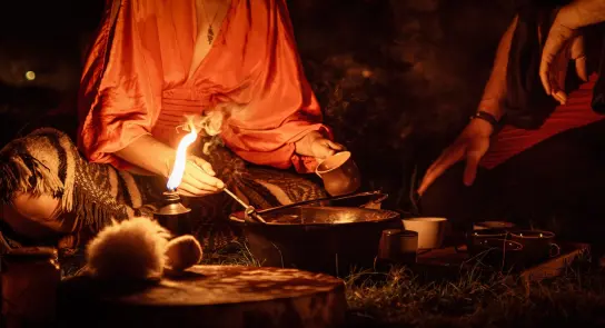 A person in a red robe pours from a pot over a flickering flame, surrounded by a cozy, dimly lit outdoor setting.