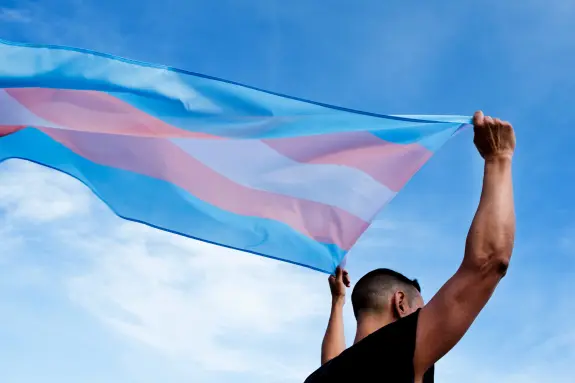 A person holds a transgender pride flag against a bright blue sky, symbolizing support and identity.
