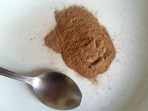 Brown spice powder arranged in a small mound on a white plate, with a silver spoon resting beside it.