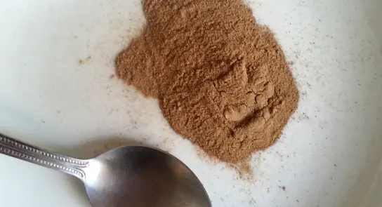 Brown spice powder arranged in a small mound on a white plate, with a silver spoon resting beside it.