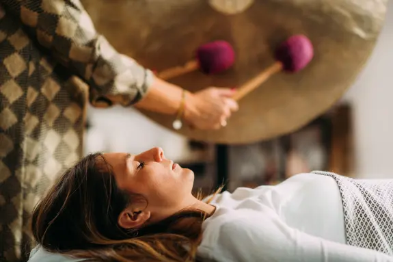 A woman lies on a table, eyes closed, as a person strikes a large gong nearby with purple mallets.