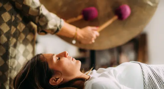 A woman lies on a table, eyes closed, as a person strikes a large gong nearby with purple mallets.