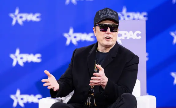 Elon Musk speaks at CPAC, wearing sunglasses and a black cap, gesturing while seated on a white chair against a blue backd...
