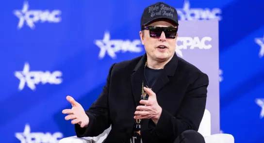 Elon Musk speaks at CPAC, wearing sunglasses and a black cap, gesturing while seated on a white chair against a blue backd...