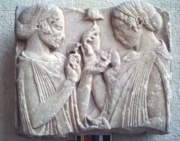 Two ancient Greek women in draped garments, holding objects, engaged in conversation, carved in relief on stone.