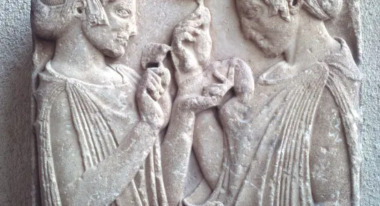 Two ancient Greek women in draped garments, holding objects, engaged in conversation, carved in relief on stone.