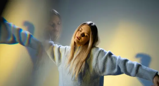 A woman with long hair in a light blue sweater, arms outstretched, against a softly lit yellow background.