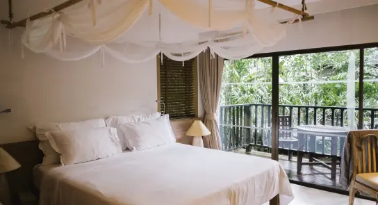Cozy bedroom with a large bed, draped canopy, and a balcony overlooking lush greenery. Soft lighting and wooden accents.