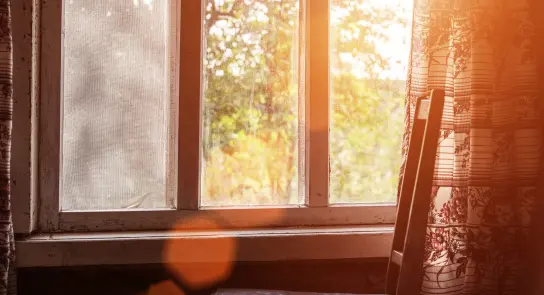 Sunlight streams through a window, illuminating a floral curtain and a wooden chair beside it.