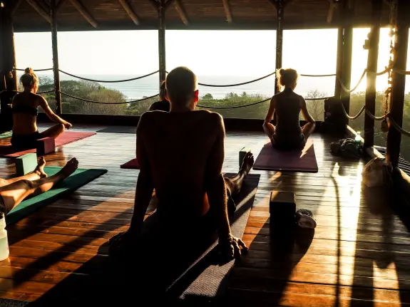 Yoga practitioners in a sunlit studio, seated on mats with a scenic ocean view in the background.