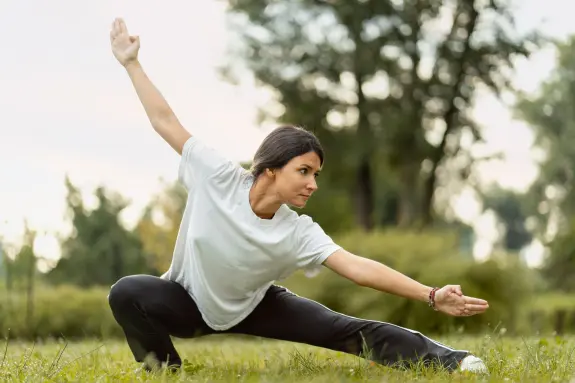 Woman in a white shirt and black pants performing a yoga pose in a grassy field, surrounded by trees.
