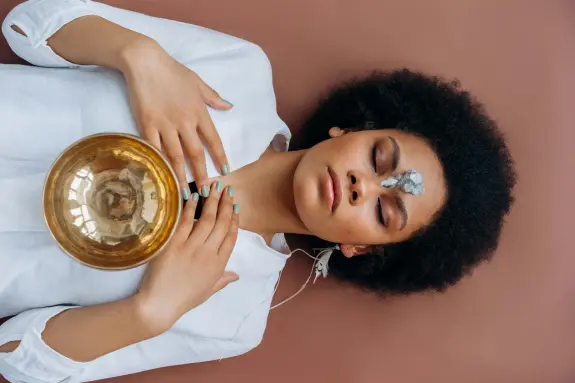 A person with curly hair lies on a brown surface, eyes closed, holding a golden bowl and wearing headphones.
