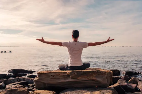 Person sitting on a rock by the water, arms outstretched, facing a calm sea under a cloudy sky.