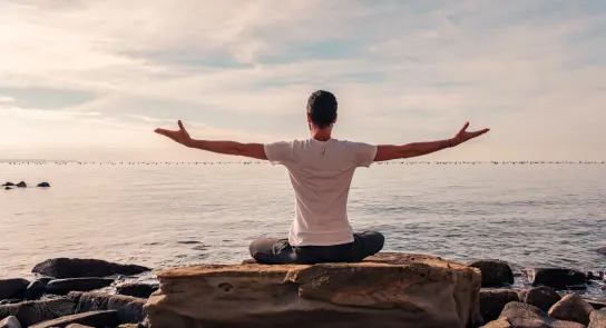 Person sitting on a rock by the water, arms outstretched, facing a calm sea under a cloudy sky.