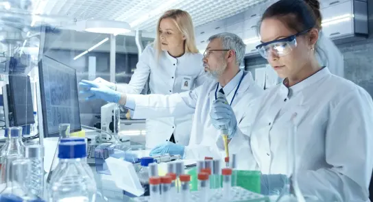 Three scientists in lab coats collaborate at a workstation, analyzing data and handling lab equipment amidst glassware.