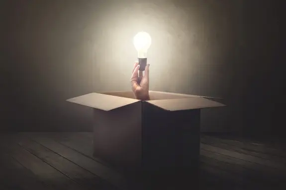 A hand emerges from a cardboard box, holding a glowing light bulb against a dimly lit background.