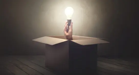 A hand emerges from a cardboard box, holding a glowing light bulb against a dimly lit background.