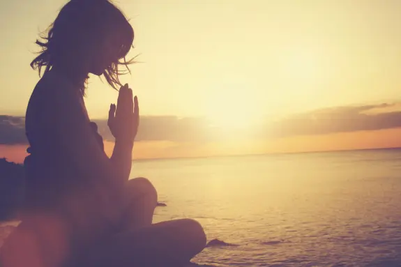 Silhouette of a woman in a prayer pose by the ocean at sunset, with warm golden hues reflecting on the water.