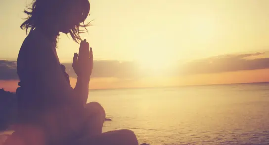 Silhouette of a woman in a prayer pose by the ocean at sunset, with warm golden hues reflecting on the water.