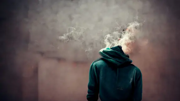 A person in a green hoodie stands with their back to the camera, surrounded by swirling smoke.