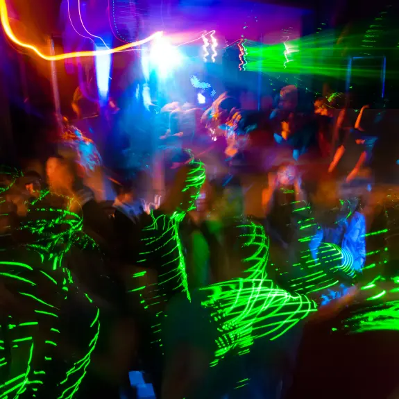 A vibrant dance floor illuminated by colorful lights and laser beams, with blurred figures moving energetically.