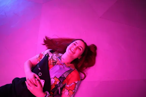 Young woman with long hair lies on a pink floor, smiling while holding a phone, wearing a colorful patterned shirt.