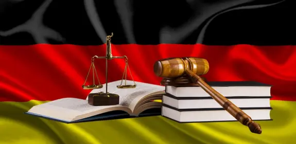 German flag backdrop with a gavel, scales of justice, and stacked law books in the foreground.