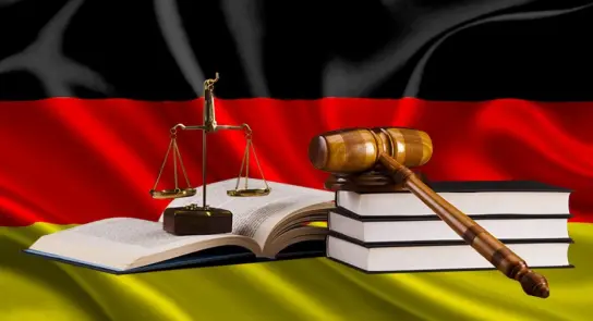 German flag backdrop with a gavel, scales of justice, and stacked law books in the foreground.