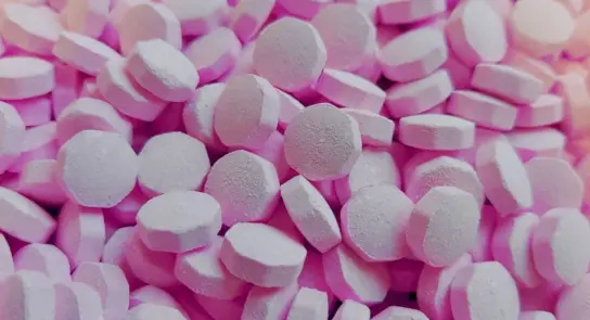 Scattered pink tablets with a round, flat shape, creating a textured surface against a light background.