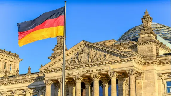German flag waving in front of the Reichstag building, showcasing its ornate architecture against a clear blue sky.