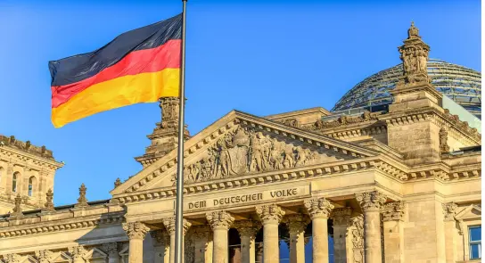 German flag waving in front of the Reichstag building, showcasing its ornate architecture against a clear blue sky.