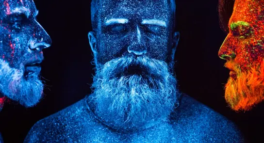 Three men with glittering, colorful body paint in a dark setting, showcasing vibrant blue, orange, and red hues.