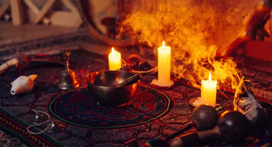 Candles glow softly around a singing bowl, surrounded by musical instruments and incense smoke on a vibrant patterned cloth.
