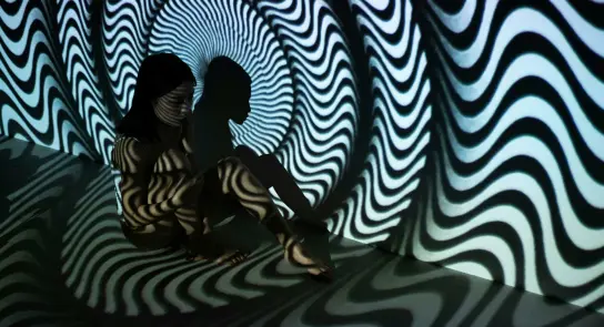 A person sits against a swirling black-and-white patterned backdrop, creating a hypnotic visual effect.