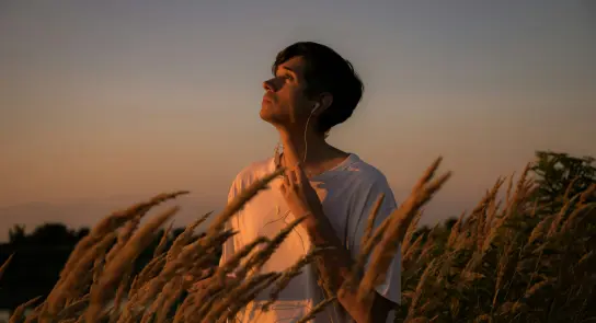 Young man in a white shirt stands among tall grass, gazing upward at a sunset sky, wearing earbuds.