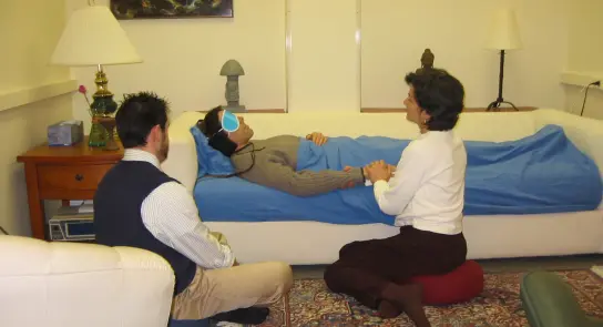 A woman lying on a couch with headphones, while a seated man and a woman kneel beside her, offering support in a calm room.