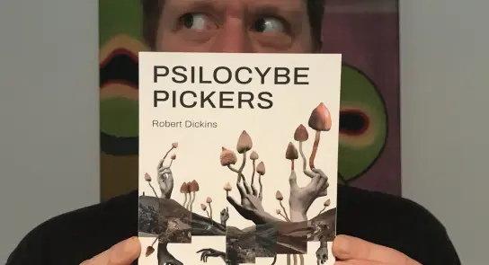 A person holds the book "Psilocybe Pickers" featuring mushrooms and figures, with abstract art in the background.