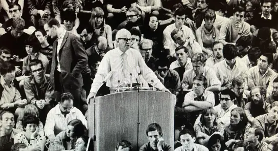 A speaker addresses a large, attentive crowd seated on the ground, with diverse faces and expressions visible.