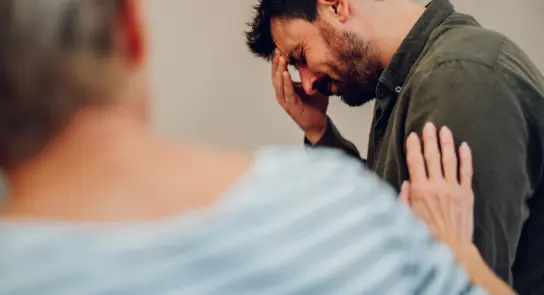 A man with a pained expression holds his forehead, while a woman gently touches his shoulder in a supportive gesture.