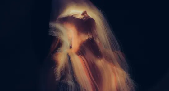 A woman with flowing hair tilts her head back, illuminated by soft, ethereal light against a dark background.