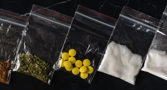 Five clear plastic bags containing various substances: spices, herbs, yellow pills, and white powder on a dark surface.