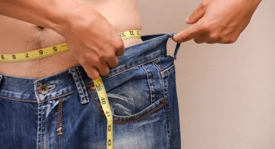 Person measuring the waist of loose-fitting jeans with a tape measure, showcasing weight loss progress.