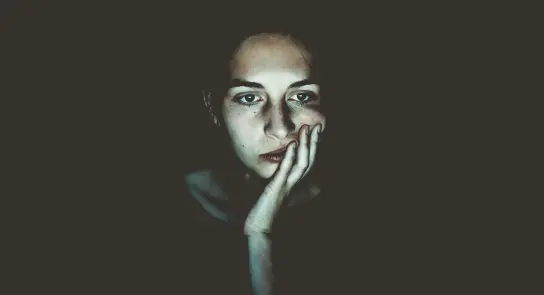 A woman with a contemplative expression rests her chin on her hand, illuminated against a dark background.