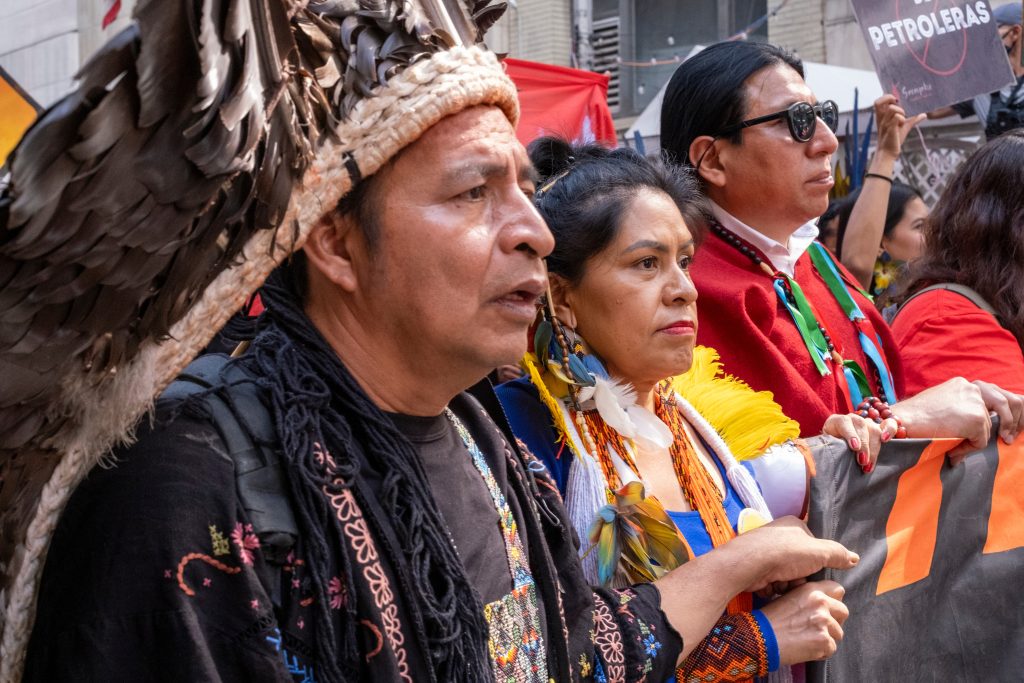 Indigenous activists in traditional attire, holding a banner, participate in a vibrant protest with colorful decorations.