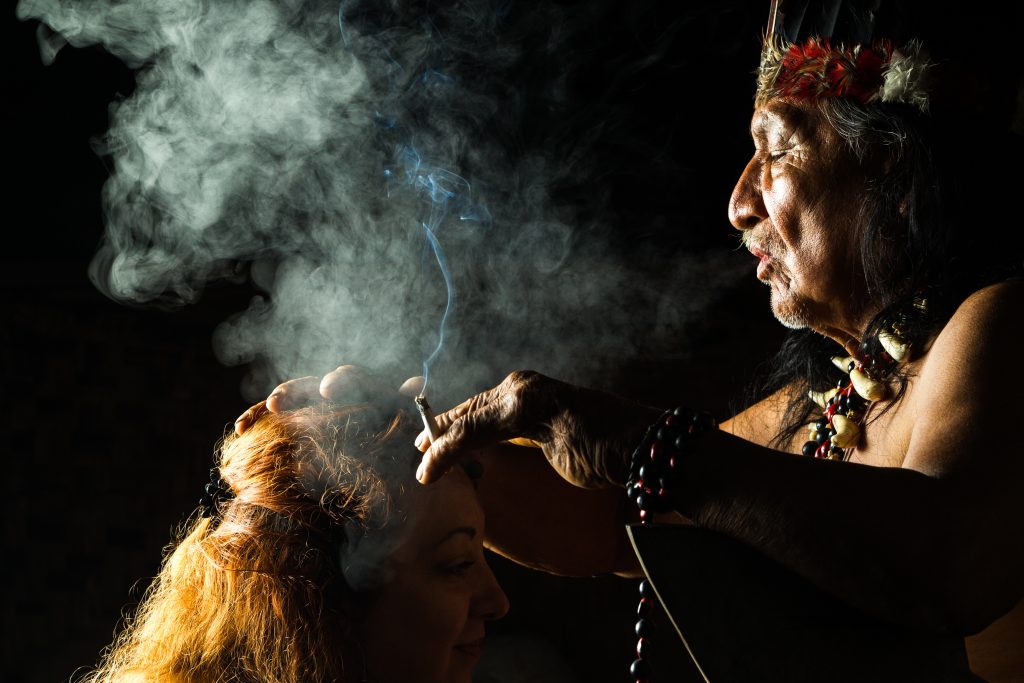 An indigenous elder performs a cleansing ritual, smoke swirling around as he touches the head of a woman.