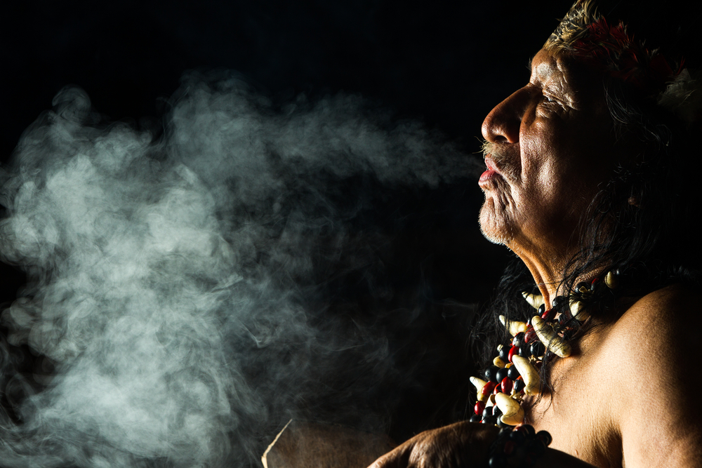 An indigenous man exhales smoke, adorned with a necklace and traditional headpiece, against a dark background.