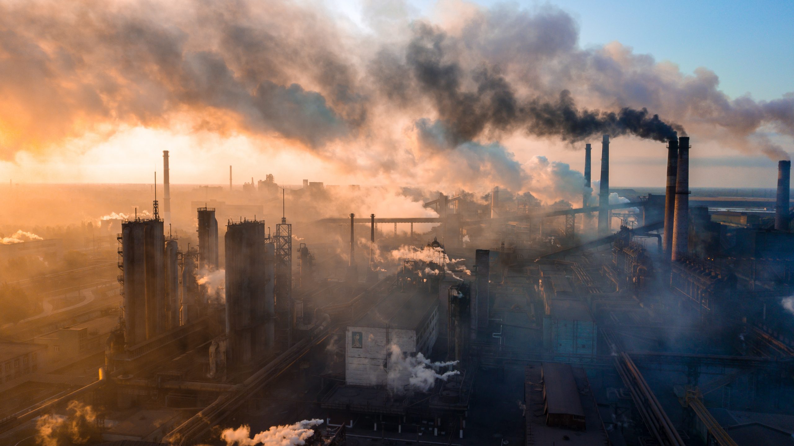 Industrial landscape with smokestacks emitting dark smoke against a hazy sunset sky.